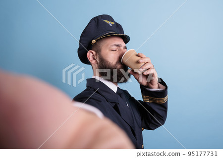 Pilot drinking coffee to go first person view, taking selfie on front camera, fpv. Middle aged airlane captain in uniform holding take away tea paper cup pox, making photo on mobile phone 95177731