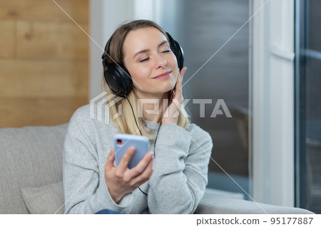 Portrait of a young beautiful woman listening to music at home in headphones from the phone, listening to audiobooks, podcasts, smiling, looking at the camera 95177887