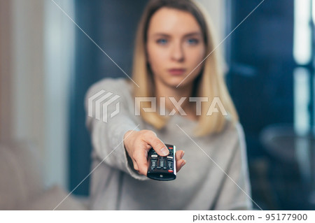 Close-up photo, portrait of a young girl holding a remote control and turning on the TV at home, in the office, hotel, looking at the camera Close-up photo, portrait of a young girl holding a remote control and turning on the TV at home, in the office, hotel, looking at the camera 95177900