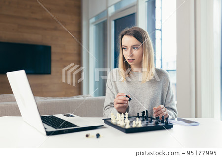 Portrait of young beautiful woman playing chess online via laptop at home, studying, thinking, competition, quarantine Portrait of young beautiful woman playing chess online via laptop at home, studying, thinking, competition, quarantine 95177995