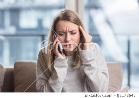Young woman sitting at home on the couch with a severe headache, trying to call a doctor with a phone, and find a solution to the problem in an online consultation 95178643