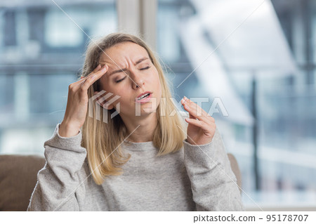 The young woman has a headache, holds her head with her hands and shows pain at home, in the office at work 95178707