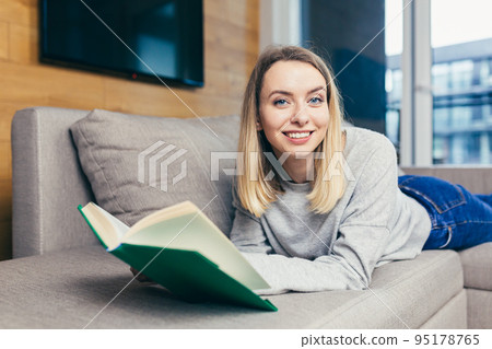 Portrait of a young beautiful woman reading a book, lying on the couch, resting, looking at the camera, smiling 95178765