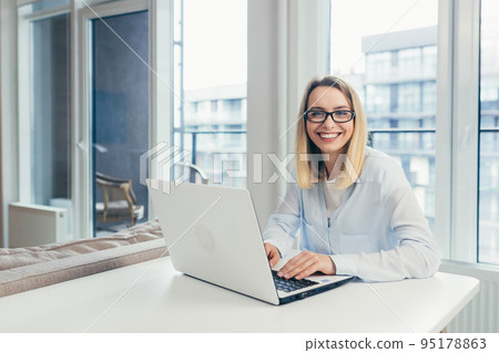 Beautiful young woman with glasses working at home on laptop, online, studying, teaching, making a video call, student, looking at camera, smiling 95178863