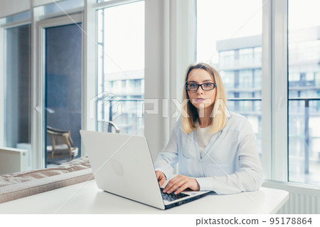 Beautiful young woman with glasses working at home on laptop, online, studying, teaching, making a video call, student, looking at camera, smiling Beautiful young woman with glasses working at home on laptop, online, studying, teaching, making a video call, student, looking at camera, smiling 95178864