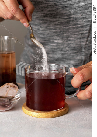 Sugar addiction. Pouring sugar with spoon in cup of tea. Black tea in glass cup. Lifestyle, action drink photo 95178894