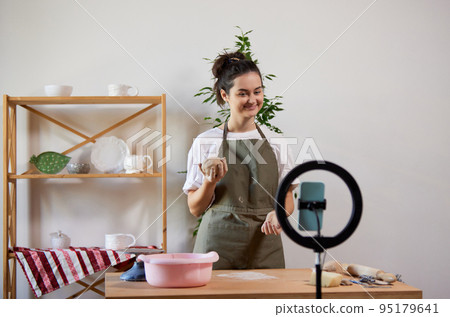Charming smiling girl, pottery master with supplies recording vlog at home studio. Hobbies and leisure activities. Remote training, online classes 95179641