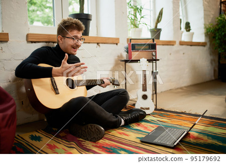Online music class. Young man siiting on floor at home and leaning to play guitar. Hobbies and leisure activities. Remote training, online classes Online music class. Young man siiting on floor at home and leaning to play guitar. Hobbies and leisure activities. Remote training, online classes 95179692