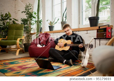 Young man siiting on floor at home and leaning to play guitar. Hobbies and leisure activities. Remote training, online classes 95179693