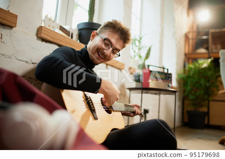 Young man siiting on floor at home and leaning to play guitar. Hobbies and leisure activities. Remote training, online classes Young man siiting on floor at home and leaning to play guitar. Hobbies and leisure activities. Remote training, online classes 95179698