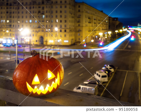 Halloween pumpkin on the background of the night city. Buildings and streaks of light from cars passing along the road. Halloween pumpkin on the background of the night city. Buildings and streaks of light from cars passing along the road. 95180058
