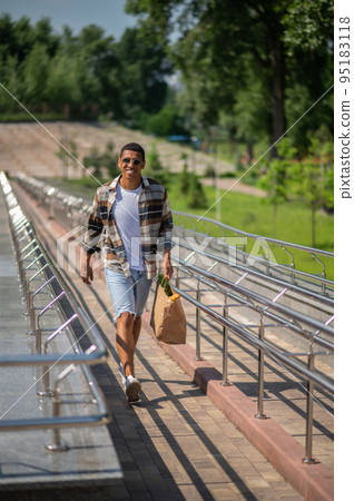 Young dark-skinned man in plaid shirt walking with a paper bag in hands Young dark-skinned man in plaid shirt walking with a paper bag in hands 95183118