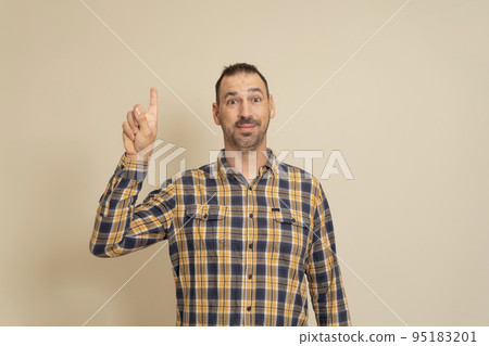 Smiling attractive man in plaid shirt posing isolated on beige wall background, studio portrait. People candid emotions lifestyle concept. Mock up copy space. Pointing the index finger up Smiling attractive man in plaid shirt posing isolated on beige wall background, studio portrait. People candid emotions lifestyle concept. Mock up copy space. Pointing the index finger up 95183201
