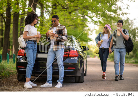 Group of riends having lunch and discussing something while standing near the car in the forest Group of riends having lunch and discussing something while standing near the car in the forest 95183945
