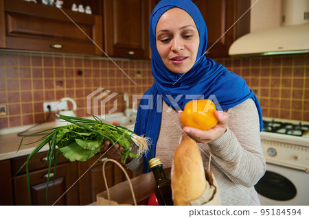 Happy young Muslim Middle-Eastern housewife in a blue hijab, lays out purchases in her cozy wooden home kitchen. Beautiful woman in good mood is sorting vegetables and fresh greens. Food purchasing 95184594