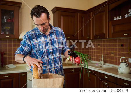 Dark-haired European man in blue checkered shirt, lays out purchases in the kitchen, sorting vegetables and greens while unpacking grocery shopping bag after shopping for groceries 95184599