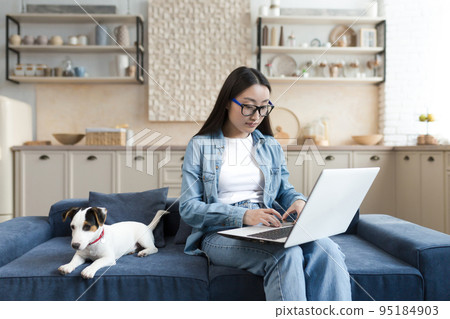 Young beautiful asian freelancer working from home remotely, woman sitting on sofa with pet small dog, jack russell terrier breed, using laptop. Young beautiful asian freelancer working from home remotely, woman sitting on sofa with pet small dog, jack russell terrier breed, using laptop. 95184903