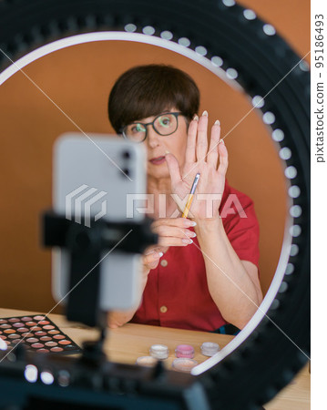 Middle-aged female beauty blogger or influencer with makeup product recording video in room at home. Using ring lamp and smartphone Middle-aged female beauty blogger or influencer with makeup product recording video in room at home. Using ring lamp and smartphone 95186493