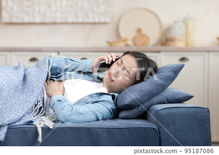 A sick Asian woman at home is lying on the sofa under a blanket, she has a severe stomach ache, the woman calls the doctor and asks for help. 95187086