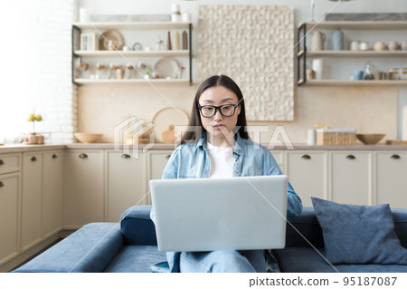 Young asian businesswoman working remotely from home sitting on sofa serious and focused using laptop for online freelance work. 95187087