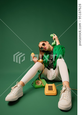 Creative portrait of young without emotions girl in green jacket sitting on chair with retro phone isolated over green background. Vintage, retro style 95187628