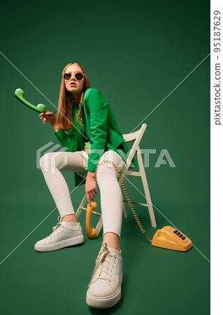 Call your parents. Creative portrait of young without emotions girl in green jacket sitting on chair with retro phone isolated over green background. 95187629