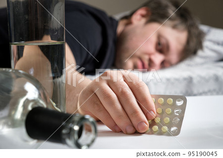 A man lies on a bed after an alcohol party, holding hangover pills in his hand 95190045