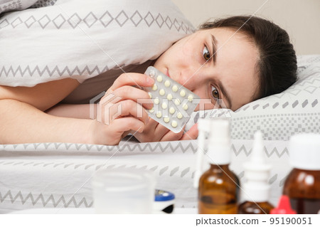 A sick woman lies in bed and holds pills in her hand. Treatment of viral and bacterial diseases at home 95190051