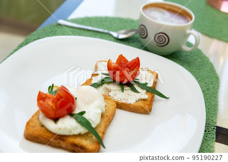 Breakfast with cup of coffee and whole wheat toasted bread with poached egg, tomato, arugula and cheese on white plate. 95190227