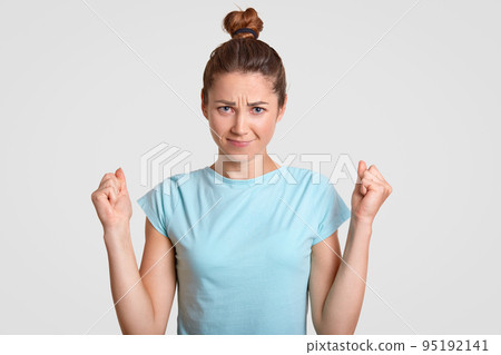 Self assured attractive woman has serious expression, keeps fists clenched, wears casual t shirt, strong desire for something, isolated over white studio wall. People, cheer, body language concept 95192141