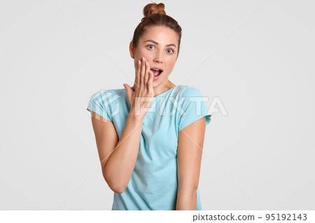 Photo of amazed fit girl keeps jaw dropped, touches cheek with hand, has bun knot, dressed in comfortable light blue t shirt, wonders latest news, isolatedd over white studio wall. Emotions concept 95192143