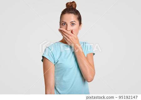 Horizontal shot of attractive shocked young woman covers mouth with palm, dressed in casual blue t shirt, tries be speechless, poses against white background, expresses wonderment. Omg concept Horizontal shot of attractive shocked young woman covers mouth with palm, dressed in casual blue t shirt, tries be speechless, poses against white background, expresses wonderment. Omg concept 95192147