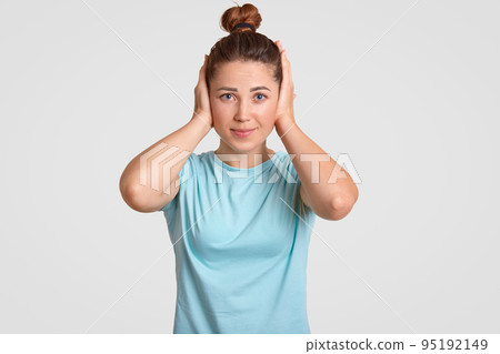 Desperate pretty woman with hair knot keeps hands on ears, ignores loud sound, looks directly at camera, dressed in casual t shirt, poses against white background. People and feelings concept Desperate pretty woman with hair knot keeps hands on ears, ignores loud sound, looks directly at camera, dressed in casual t shirt, poses against white background. People and feelings concept 95192149