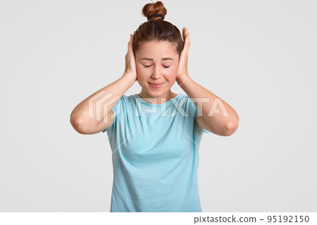 Young female model covers ears with both palms, dressed in casual t shirt, avoids loud sound, has dark hair combed in knot, isolated over white background. Cute girl ignores listening something Young female model covers ears with both palms, dressed in casual t shirt, avoids loud sound, has dark hair combed in knot, isolated over white background. Cute girl ignores listening something 95192150