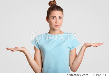 Hesitant young woman shrugs shoulders with uncertainy, cant make decision, clasps palms, looks clueless at camera, dressed in blue t shirt, isolated over white background. So, what to do now? 95192151