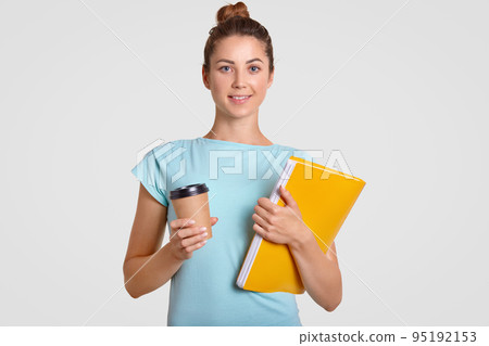 Shot of pleasant looking woman with hair bun has delighted expression, holds takeaway coffee and yellow folder, dressed casually, rests after having classes, isolated over white studio background Shot of pleasant looking woman with hair bun has delighted expression, holds takeaway coffee and yellow folder, dressed casually, rests after having classes, isolated over white studio background 95192153