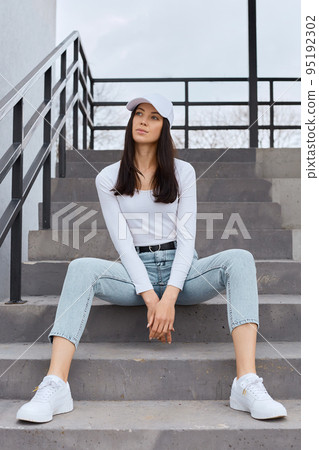 Beautiful young adult woman wearing jeans, white shirt and baseball cap sitting on stairs outdoors and looking away, having serious facial expression, dreams or thinks about important things. 95192302