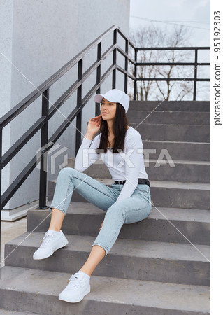 Attractive female wearing stylish jeans, white shirt and baseball cap, sitting on stairs and looking away with thoughtful expression, posing outdoor near building. 95192303