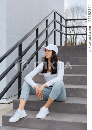 Full length photo of dark haired female wearing jeans, white shirt and baseball cap, posing outdoors while sitting on stairs near building, looking away with confident facial expression. 95192304