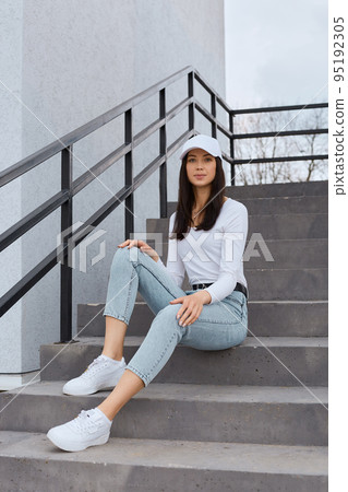 Winsome Caucasian woman sits on stairs near wall, keeping hands on knees, wearing jeans, white shirt and baseball cap, looking at camera, expressing confidence. 95192305