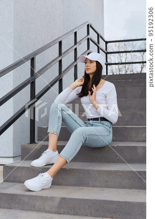 Young attractive woman with perfect body dresses jeans, white shirt and baseball cap, posing outdoors on stairs, looking away with thoughtful expression, touching her beautiful hair. 95192306