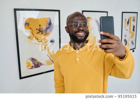 African young man in eyeglasses using his mobile phone for streaming standing at art gallery 95195015