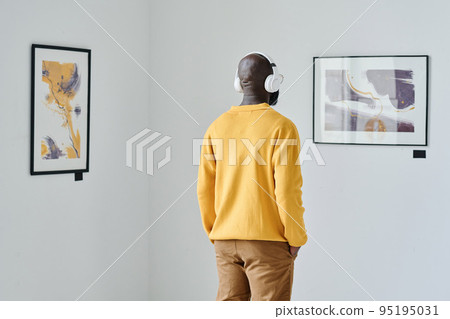 Rear view of African young man in wireless headphones looking at modern art and listening to guide Rear view of African young man in wireless headphones looking at modern art and listening to guide 95195031