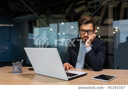 sad and depressed businessman in depression working inside modern office building, man in business suit with glasses and beard using laptop for work, boss disappointed with work result. 95195167