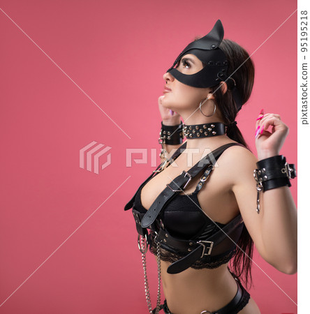 brunette in bdsm leather straps on a pink background in a cat mask in handcuffs for sex games 95195218