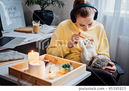 Relaxed young woman with cat sitting near table with burning candles and aroma sticks at home and do meditation and relaxation 95195823