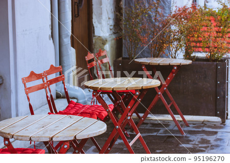 Classic empty cafe restaurant terrace after rain without people. Folding chairs with red soft cushions, wooden round tables. A patio of a bar. Garden furniture. Nice area for relaxing on a city street Classic empty cafe restaurant terrace after rain without people. Folding chairs with red soft cushions, wooden round tables. A patio of a bar. Garden furniture. Nice area for relaxing on a city street 95196270