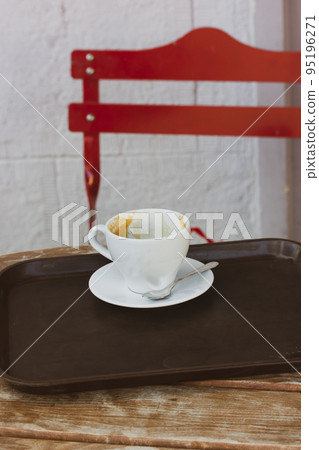 A white coffee cup on a saucer stands on a tray on a wooden table on the terrace of a cafe, a coffee shop in an urban urban environment. Conceptual vertical photo with a hot beverage drink. Red chair. A white coffee cup on a saucer stands on a tray on a wooden table on the terrace of a cafe, a coffee shop in an urban urban environment. Conceptual vertical photo with a hot beverage drink. Red chair. 95196271