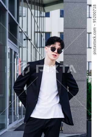 handsome young man in a black shirt standing against the backdrop of city buildings  and looking away through black sunglasses 95200718
