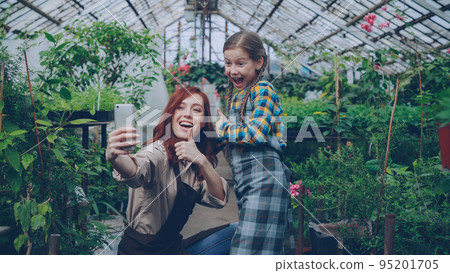 Loving mother and playful little daughter are taking selfie in greenhouse posing, gesturing and making funny faces. Family, green plants and modern technology concept. 95201705